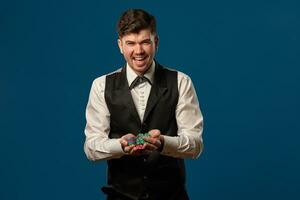 Newbie in poker, in black vest and trousers, white shirt. Holding some colored chips. Posing against blue background. Gambling, casino. Close-up. photo