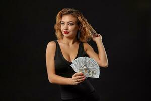 Ginger girl in dark dress posing holding fan of hundred-dollar bills in her hands standing against a black studio background. Casino, poker. Close-up. photo