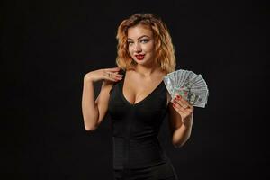 Ginger girl in dark dress posing holding fan of hundred-dollar bills in her hands standing against a black studio background. Casino, poker. Close-up. photo