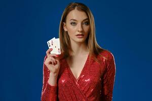 Blonde woman with make-up, in red sequin dress is showing two aces, posing on blue background. Gambling, poker, casino. Close-up. photo