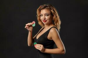 Ginger girl wearing dark dress is posing holding red and green chips in her hands standing against black studio background. Casino, poker. Close-up. photo