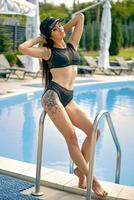 Portrait of a woman having rest and posing near a swimming pool. Dressed in a black swimsuit and sun visor. photo