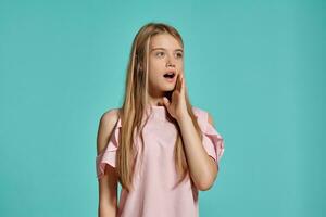 estudio retrato de un hermosa niña rubia adolescente en un rosado camiseta posando terminado un azul antecedentes. foto