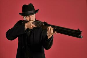 Middle-aged man with beard, mustache, in black jacket and hat, holding a gun while posing against a red background. Sincere emotions concept. photo