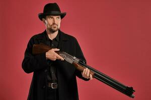 Middle-aged man with beard, mustache, in black jacket and hat, holding a gun while posing against a red background. Sincere emotions concept. photo