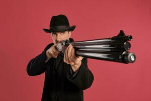 Middle-aged man with beard, mustache, in black jacket and hat, holding a gun while posing against a red background. Sincere emotions concept. photo