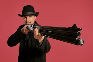 Middle-aged man with beard, mustache, in black jacket and hat, holding a gun while posing against a red background. Sincere emotions concept. photo