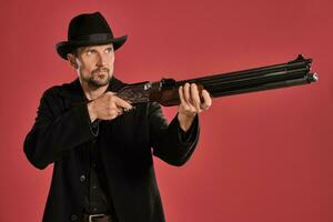 Middle-aged man with beard, mustache, in black jacket and hat, holding a gun while posing against a red background. Sincere emotions concept. photo