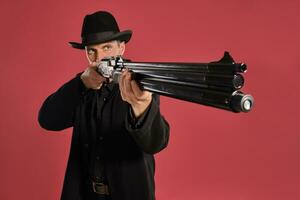 Middle-aged man with beard, mustache, in black jacket and hat, holding a gun while posing against a red background. Sincere emotions concept. photo