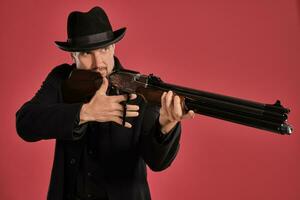 Middle-aged man with beard, mustache, in black jacket and hat, holding a gun while posing against a red background. Sincere emotions concept. photo