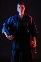 Kendo fighter wearing in an armor, traditional kimono is holding his helmet and shinai bamboo sword while posing on a black background. Close up. photo