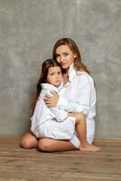 Indoor portrait of a beautiful mother with her charming little daughter posing against a gray wall. photo