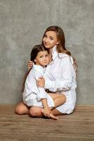 Indoor portrait of a beautiful mother with her charming little daughter posing against a gray wall. photo