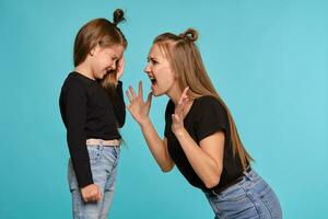 Mom and daughter with a funny hairstyles, dressed in black shirts and blue denim jeans are posing against a blue studio background. Close-up shot. photo