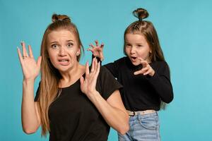 Mom and daughter with a funny hairstyles, dressed in black shirts and blue denim jeans are posing against a blue studio background. Close-up shot. photo