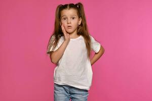 Little girl with ponytails, dressed in white t-shirt and blue jeans is posing against a pink studio background. Close-up shot. Sincere emotions. photo