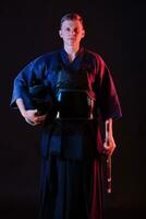 Kendo fighter wearing in an armor, traditional kimono is holding his helmet and shinai bamboo sword while posing on a black background. Close up. photo