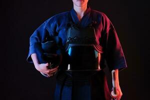 Kendo fighter wearing in an armor, traditional kimono is holding his helmet and shinai bamboo sword while posing on a black background. Close up. photo