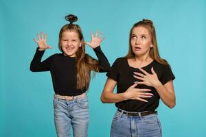 Mom and daughter with a funny hairstyles, dressed in black shirts and blue denim jeans are posing against a blue studio background. Close-up shot. photo