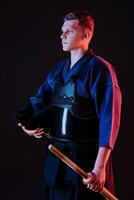 Kendo fighter wearing in an armor, traditional kimono is holding his helmet and shinai bamboo sword while posing on a black background. Close up. photo