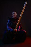 Close up. Kendo fighter wearing in an armor, traditional kimono, helmet, sitting, practicing martial art with shinai bamboo sword, black background. photo