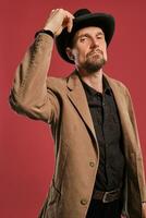 Middle-aged man with beard and mustache, wears black hat and brown jacket posing against a red background. Sincere emotions concept. photo