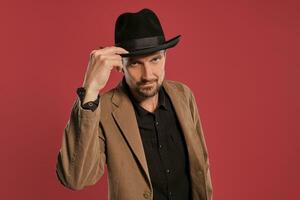 Middle-aged man with beard and mustache, wears black hat and brown jacket posing against a red background. Sincere emotions concept. photo