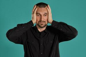 Middle-aged man with beard and mustache, wears black shirt, posing against a blue background. Sincere emotions concept. photo