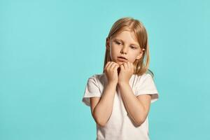 de cerca estudio Disparo de un encantador rubia pequeño niña en un blanco camiseta posando en contra un azul antecedentes. foto