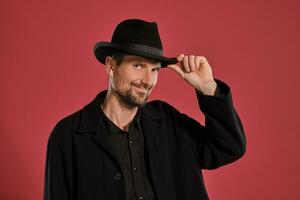 Middle-aged man with beard and mustache, wears black hat and jacket posing against a red background. Sincere emotions concept. photo