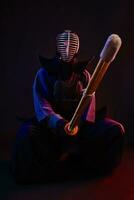 Close up. Kendo fighter wearing in an armor, traditional kimono, helmet, sitting, practicing martial art with shinai bamboo sword, black background. photo