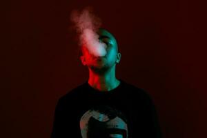 Close-up studio shot of a young bald guy vaping, blowing out a cloud of smoke on red background. photo