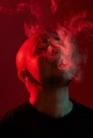 Close-up studio shot of a young bald guy vaping, blowing out a cloud of smoke on red background. photo