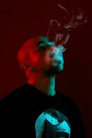 Close-up studio shot of a young bald guy vaping, blowing out a cloud of smoke on red background. photo