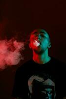 Close-up studio shot of a young bald guy vaping, blowing out a cloud of smoke on red background. photo