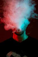 Close-up studio shot of a young bald guy vaping, blowing out a cloud of smoke on red background. photo