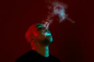 Close-up studio shot of a young bald guy vaping, blowing out a cloud of smoke on red background. photo