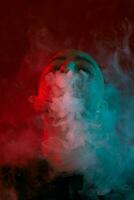 Close-up studio shot of a young bald guy vaping, blowing out a cloud of smoke on red background. photo