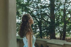 Beautiful curly hair young woman in light blue simple dress on balcony, park trees background photo