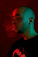 Close-up studio shot of a young bald guy vaping, blowing out a cloud of smoke on red background. photo