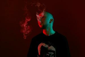 Close-up studio shot of a young bald guy vaping, blowing out a cloud of smoke on red background. photo