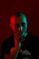 Close-up studio shot of a young bald guy vaping, blowing out a cloud of smoke on red background. photo