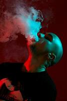 Close-up studio shot of a young bald guy vaping, blowing out a cloud of smoke on red background. photo