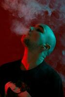 Close-up studio shot of a young bald guy vaping, blowing out a cloud of smoke on red background. photo