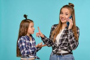 Mom and daughter dressed in checkered shirts and blue denim jeans are using smartphone while posing against a blue studio background. Close-up shot. photo