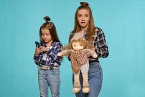 mamá y hija vestido en a cuadros camisas y azul mezclilla pantalones son utilizando teléfono inteligente mientras posando en contra un azul estudio antecedentes. de cerca disparo. foto