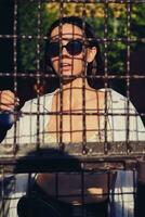 Portrait of girl in sunglasses posing in city behind a trellised fence. Dressed in top with floral print, white shirt, black trousers, waist bag. photo