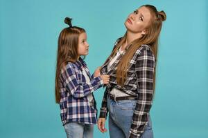 Mom and daughter with a funny hairstyles, dressed checkered shirts and blue denim jeans are posing against a blue studio background. Close-up shot. photo