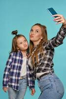 Mom and daughter dressed in checkered shirts and blue denim jeans are making selfie while posing against a blue studio background. Close-up shot. photo