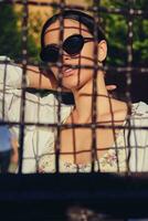 Portrait of girl in sunglasses posing in city behind a trellised fence. Dressed in top with floral print, white shirt, black trousers, waist bag. photo